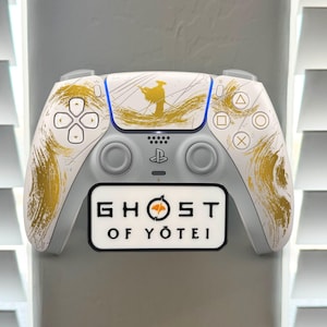 May include: A white and gray PlayStation 5 controller with gold brushstroke accents and a blue stripe. The controller has the text "GHOST OF YOTEI" on a black and white label.