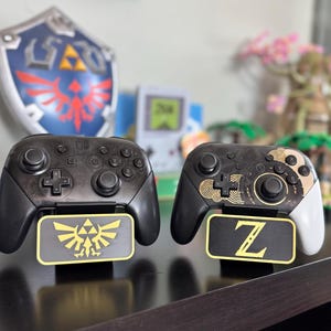 May include: Two black and white Nintendo Switch controllers with custom faceplates. The left controller has a gold Triforce symbol and the right controller has a gold Z symbol.
