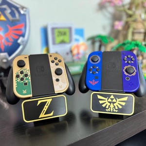 May include: Two Nintendo Switch controllers with custom designs. The left controller is gold with a green and black design. The right controller is blue with a gold design. Both controllers are on stands with a gold Zelda logo.