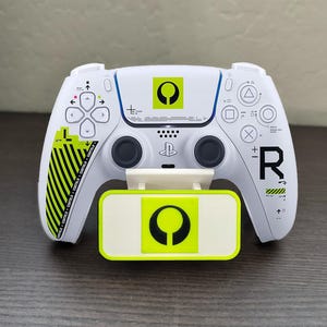 May include: White gaming controller with black buttons and a matching charging stand. The controller features black and neon green accents, with the letter "R" and other symbols. The text "CONTIER CONTRER CONTOUR CONTRER CONTR" is printed on the left side.