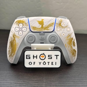 May include: A white and gray game controller with gold brushstroke accents and a blue stripe. The controller has a white stand with the text "GHOST OF YŌTEI" in black. The controller has the PlayStation logo.