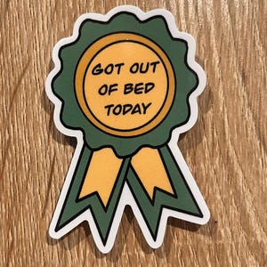 May include: A sticker shaped like a green and yellow award ribbon with a circular center. The center features the text "GOT OUT OF BED TODAY" in black capital letters. The design includes a scalloped edge and two downward-pointing ribbon tails.