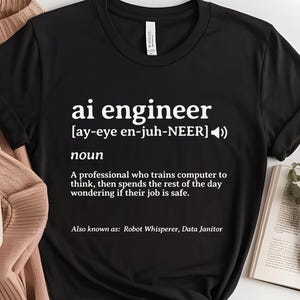 May include: Black t-shirt with white text defining "ai engineer" as a noun, including pronunciation and a humorous description. The shirt also lists "Robot Whisperer, Data Janitor" as alternative titles.