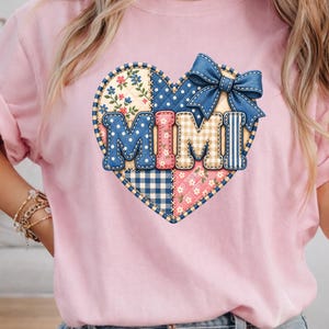 May include: A light pink t-shirt featuring a large heart-shaped graphic on the chest. The heart is designed with a patchwork pattern of blue, pink, and beige fabrics, including floral and gingham prints. The word MIMI is centered in the heart.