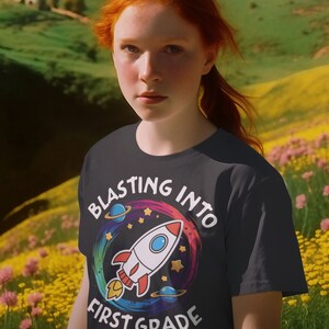 Op de afbeelding: Een zwart T-shirt met een kleurrijke afbeelding van een raket die de ruimte in schiet. De afbeelding bevat de tekst "Blasting into First Grade".