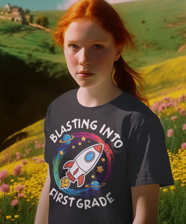 Op de afbeelding: Een zwart T-shirt met een kleurrijke afbeelding van een raket die de ruimte in schiet. De afbeelding bevat de tekst "Blasting into First Grade".