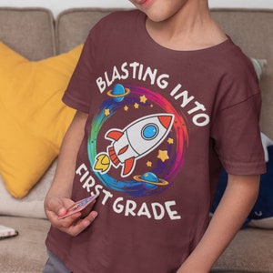 Op de afbeelding: Een maroon t-shirt met een kleurrijk raketontwerp en de tekst "Blasting into First Grade".