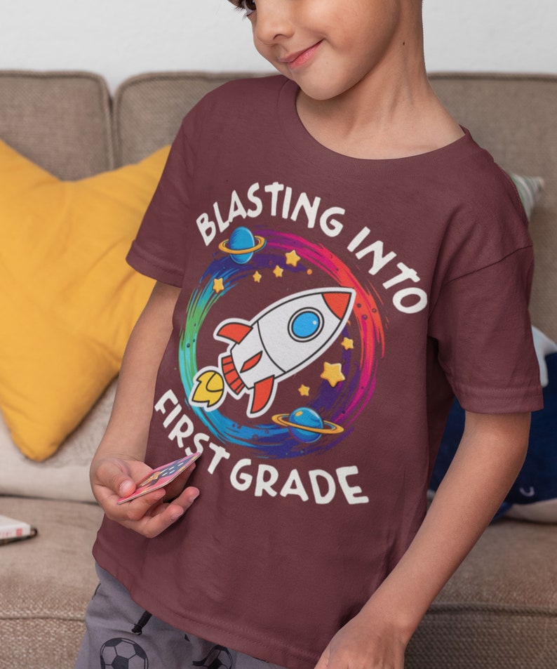 Op de afbeelding: Een maroon t-shirt met een kleurrijk raketontwerp en de tekst "Blasting into First Grade".