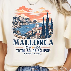 May include: A cream-colored t-shirt featuring a graphic print of a coastal landscape in Mallorca with a total solar eclipse in the sky. The text reads MALLORCA, TOTAL SOLAR ECLIPSE, and AUGUST 12 2026 in dark blue and orange ink colors.
