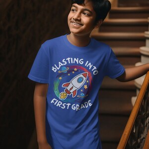 Op de afbeelding: Een blauw T-shirt met een kleurrijke raketgrafiek en de tekst "Blasting into First Grade".