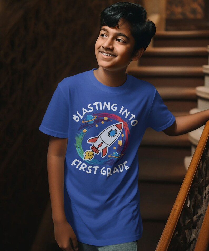 Op de afbeelding: Een blauw T-shirt met een kleurrijke raketgrafiek en de tekst "Blasting into First Grade".