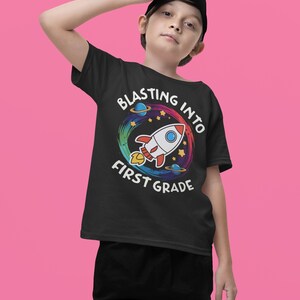 Op de afbeelding: Een zwart T-shirt met een kleurrijk raketontwerp en de tekst "Blasting into First Grade".