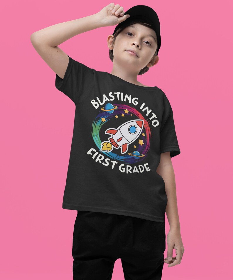 Op de afbeelding: Een zwart T-shirt met een kleurrijk raketontwerp en de tekst "Blasting into First Grade".