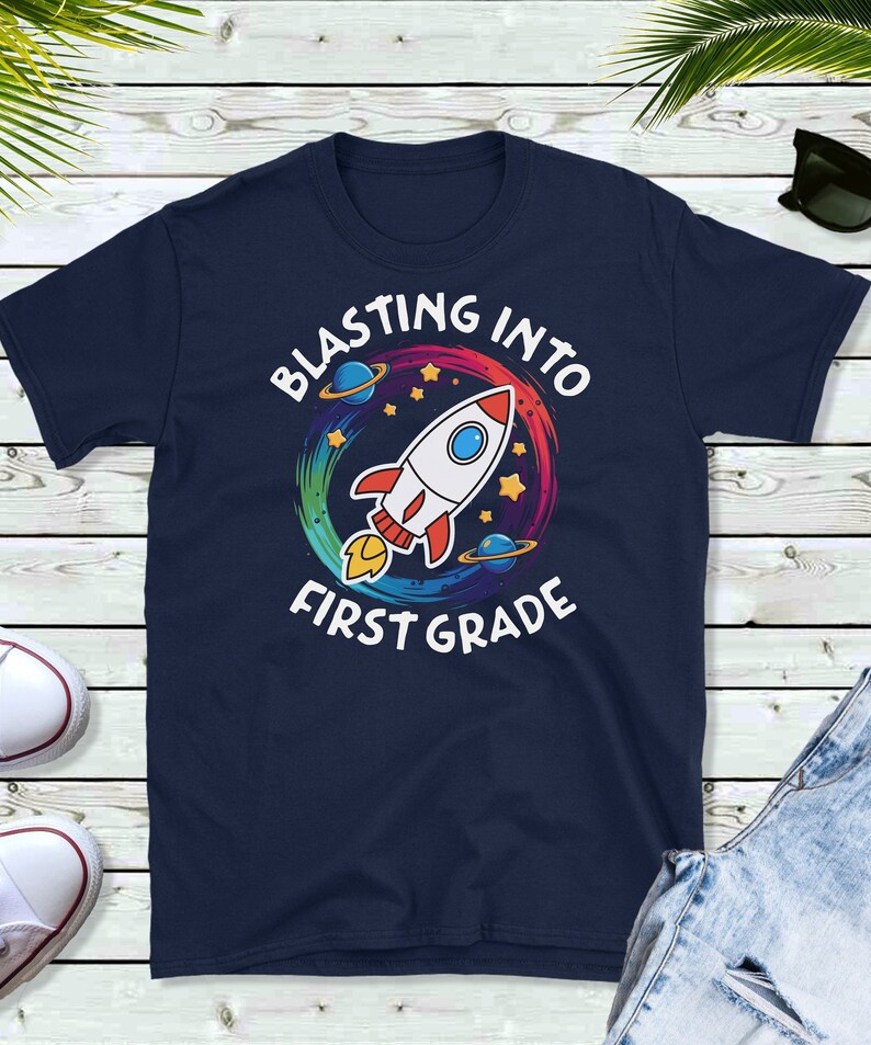 Op de afbeelding: Donkerblauw T-shirt met een kleurrijk raketontwerp en de tekst "Blasting into First Grade".