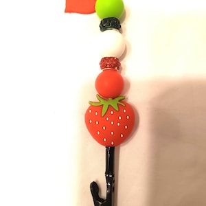 May include: A decorative lanyard clip featuring a red strawberry charm with white seeds, a red heart, and various colorful beads. The clip is black and has a lobster claw clasp.