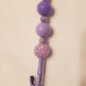 May include: A purple beaded lanyard with a lobster claw clasp. The lanyard features various sized purple beads, silver accents, and a decorative cluster of pink rhinestones. A purple clip is attached at the bottom.