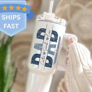 May include: White tumbler with a handle and a straw. The tumbler has the word "DAD" in navy blue, with the names "Carter Sarah Lisa" in a script font. A blue star and heart are also featured.