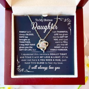 May include: A silver necklace with a sparkling pendant, presented in a gift box. The pendant is a knot design with a central gemstone. The box contains a message to a bonus daughter, expressing love and appreciation. The box is dark red.