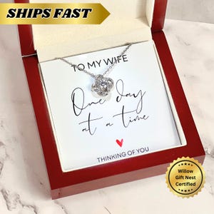 Könnte beinhalten: Eine silberfarbene Halskette mit einem Knotenanhänger, präsentiert in einer roten Holzgeschenkbox. Die Box enthält eine Karte mit der Aufschrift "TO MY WIFE, One day at a time, THINKING OF YOU." Ein goldener Aufkleber lautet "Willow Gift Nest Certified."