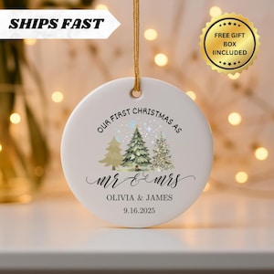May include: A white ceramic Christmas ornament with a gold string. The ornament features the text "OUR FIRST CHRISTMAS AS mr & mrs OLIVIA & JAMES 9.16.2025" and illustrations of Christmas trees. The words "SHIPS FAST" and "FREE GIFT BOX INCLUDED" are also visible.