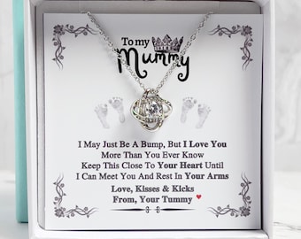 Puede incluir: Una caja de regalo blanca con un collar de plata en su interior. El collar tiene un colgante de nudo con un cristal transparente. La caja tiene un mensaje que dice: "To my Mummy I May Just Be A Bump, But I Love You More Than You Ever Know Keep This Close To Your Heart Until I Can Meet You And Rest In Your Arms Love, Kisses & Kicks From, Your Tummy".