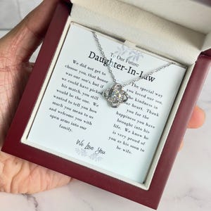 May include: A wooden box with a white card inside. The card has a message that reads "To Our Future Daughter-In-Law" and a silver necklace with a knot pendant. The necklace is in a box with a red velvet lining.