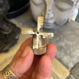 May include: A small, antique-style metal windmill with four blades. The windmill is a light gold color with a textured surface, and is held in a hand. The base has a ridged design.