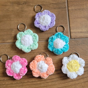 May include: Six crocheted flower keychains in various colors, including purple, green, blue, pink, orange, and yellow. Each flower has a white center and a fluffy texture.