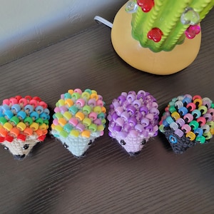 May include: Five crocheted hedgehogs with colorful beaded spines. The hedgehogs are arranged in a row on a dark brown surface. The colors of the beads are rainbow, pastel, purple, black and green.