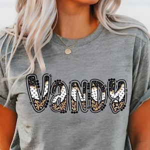 Könnte beinhalten: Ein graues T-Shirt mit dem Wort "VANDY" in einer auffälligen, dekorativen Schriftart. Die Buchstaben weisen eine Mischung aus Leopardenmuster, weißen Kreuzen und schwarzen Akzenten auf. Das T-Shirt hat kurze Ärmel.