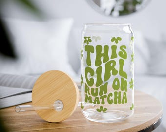 May include: A clear glass can-shaped drinking glass with the text "THIS GIRL RUN ON MATCHA" in green. The glass has a bamboo lid and a clear straw. The glass is decorated with green floral accents.