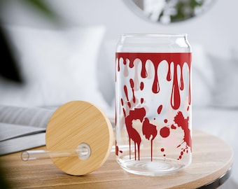 Può includere: Un bicchiere trasparente a forma di lattina con coperchio e cannuccia in bambù. Il vetro è decorato con un disegno a spruzzi di sangue rosso. Il bicchiere è appoggiato su un tavolo di legno.