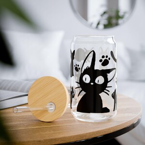 May include: A clear glass can-shaped cup with a black cat graphic and paw prints. The cup has a bamboo lid and a clear straw. The cup is sitting on a wooden table.