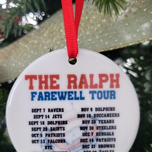 Può includere: Una palla di Natale bianca e rotonda con la scritta "THE RALPH FAREWELL TOUR" in rosso e blu. L'ornamento elenca le date e le squadre di football americano. Un nastro rosso è attaccato per appenderlo. L'ornamento è su un albero di Natale.