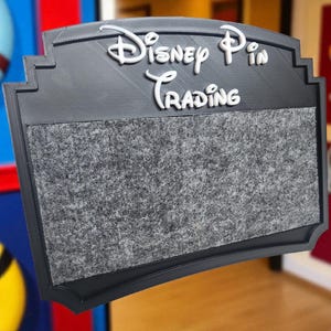 May include: A black rectangular sign with the words "Disney Pin Trading" in white, stylized lettering. The sign has a gray felt-like surface for displaying pins. The sign is framed in black and has a decorative top edge.