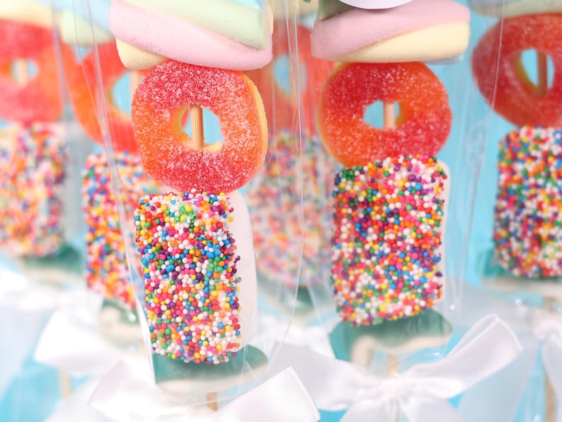 Pu&ograve; includere: Primo piano di spiedini di caramelle colorate. Ogni spiedino presenta un marshmallow, una caramella a forma di anello zuccherato e un marshmallow rettangolare ricoperto di zuccherini arcobaleno. Le leccornie sono confezionate in cellophane trasparente con fiocchi bianchi.