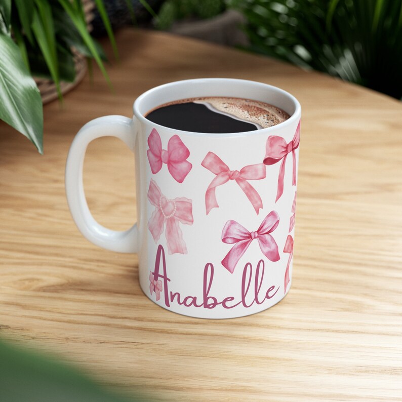 May include: White ceramic mug with a pink and white bow design and the name "Anabelle" written in pink script.