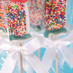 Pu&ograve; includere: Primo piano di lecca lecca colorati. Ogni lecca lecca &egrave; ricoperto di zuccherini arcobaleno e avvolto in cellophane trasparente. Nastri di raso bianco sono legati attorno ai bastoncini di legno, creando un aspetto festoso. Lo sfondo &egrave; azzurro.