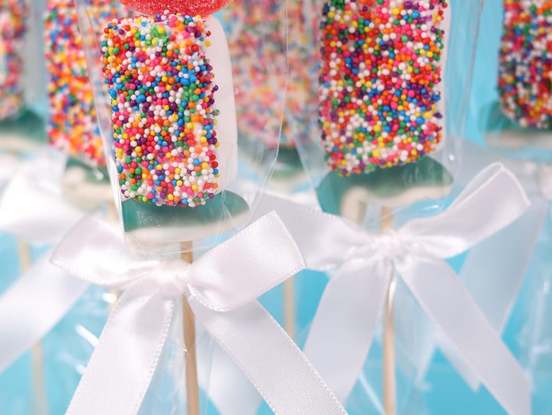 Pu&ograve; includere: Primo piano di lecca lecca colorati. Ogni lecca lecca &egrave; ricoperto di zuccherini arcobaleno e avvolto in cellophane trasparente. Nastri di raso bianco sono legati attorno ai bastoncini di legno, creando un aspetto festoso. Lo sfondo &egrave; azzurro.