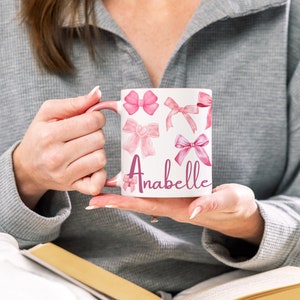 May include: White ceramic mug with a pink handle and pink watercolor bow designs. The mug has the name "Anabelle" written in pink cursive lettering.