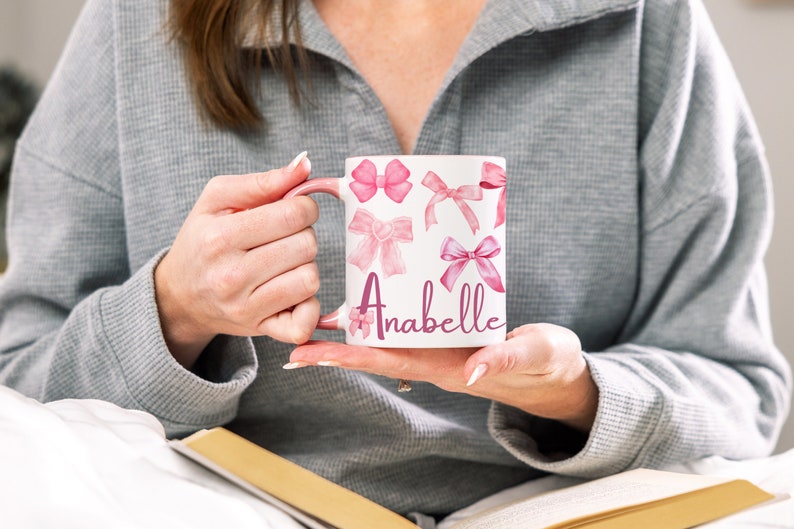 May include: White ceramic mug with a pink handle and pink watercolor bow designs. The mug has the name "Anabelle" written in pink cursive lettering.
