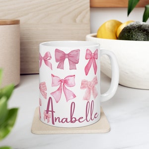 May include: White ceramic mug with a pink and white design of bows. The mug has the name "Anabelle" written in pink on the bottom.