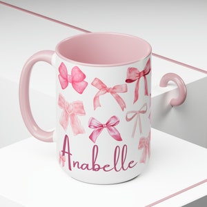 May include: A white ceramic mug with a pink handle and pink rim. The mug features a pink and white watercolor design of bows and the name "Anabelle" in pink lettering.