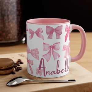 May include: A pink and white ceramic mug with a pink rim and handle. The mug features a pattern of pink watercolor bows and the name "Anabelle" in pink script.