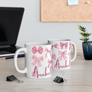 May include: Two white ceramic mugs with pink watercolor bows and the name "Anabell" printed on them.