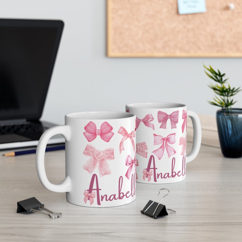 May include: Two white ceramic mugs with pink watercolor bows and the name "Anabell" printed on them.