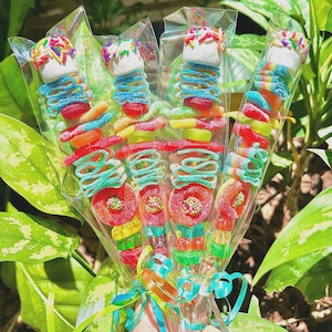 May include: A bouquet of candy skewers, each wrapped in clear cellophane. The skewers feature a variety of colorful gummy candies, including rings, worms, and marshmallows topped with sprinkles. A teal and orange ribbon secures the bouquet.