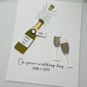 May include: A white card with a gold glitter champagne bottle and two champagne glasses. The card says "On your wedding day" and "Congrats to you both!" with the names "Joann & Cooper" below.