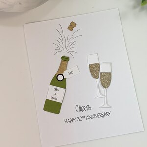 May include: A white card with a green champagne bottle, two champagne glasses, and a cork popping out of the bottle. The card says "Cheers Happy 30th Anniversary" and "Chris & Carole" on the bottle. The bottle has a tag that says "Love".