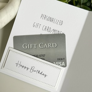 May include: A white card with the text "Personalized Gift Card/Money" and a gray gift card with the text "Gift Card" and "Debit Visa". The card has a white label with the text "Happy Birthday" on it.